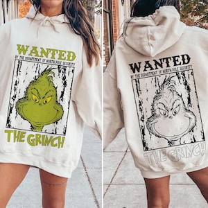 May include: Cream-colored hooded sweatshirt featuring a Grinch graphic. The front displays a green Grinch face with the text "WANTED" and "THE GRINCH". The back shows a black and white Grinch face with the same text.