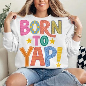 May include: White sweatshirt with the phrase "BORN TO YAP!" in colorful, playful lettering. The design features pink, orange, yellow, blue, and green text with star accents. The sweatshirt is a casual, comfortable style.