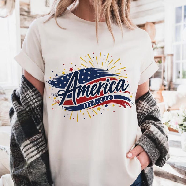 May include: A cream-coloured t-shirt with the word "America" in a decorative script, and the years "1776-2026" beneath. The design uses red, white, and blue, with stars and stripes, and yellow starbursts.
