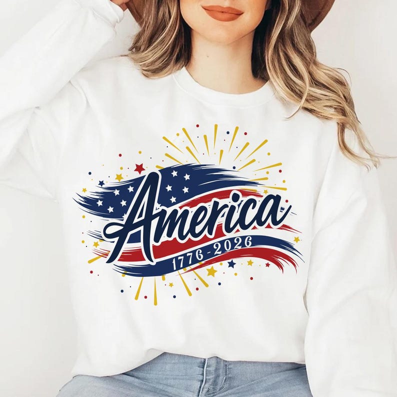 May include: White sweatshirt with the word "America" in a script font, with a design featuring the American flag colours and stars. The design includes the years "1776-2026" and gold starbursts.