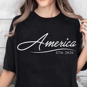 May include: Black t-shirt with the word "America" in a white script font, a star, and the dates "1776-2026" below. The shirt is a crew neck style.
