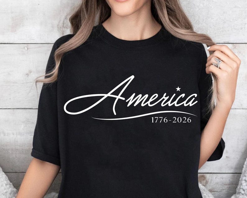 May include: Black t-shirt with the word "America" in a white script font, a star, and the dates "1776-2026" below. The shirt is a crew neck style.
