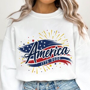 May include: White sweatshirt with a patriotic design. The graphic features the word "America" in navy blue script, with a red and blue brushstroke design and the dates "1776-2026". Gold and red starbursts and stars complete the design.