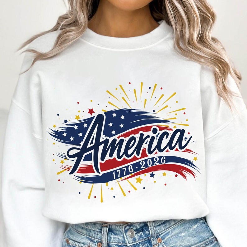 May include: White sweatshirt with a patriotic design. The graphic features the word "America" in navy blue script, with a red and blue brushstroke design and the dates "1776-2026". Gold and red starbursts and stars complete the design.