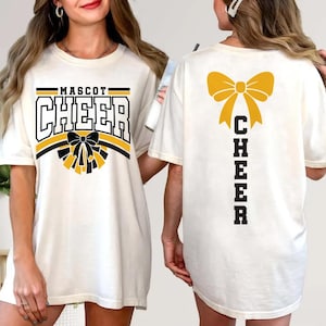 May include: Cream-colored t-shirt with the word "MASCOT CHEER" in black and gold lettering. The front features a bow and pom-pom design. The back of the shirt displays a gold bow and the word "CHEER" vertically.