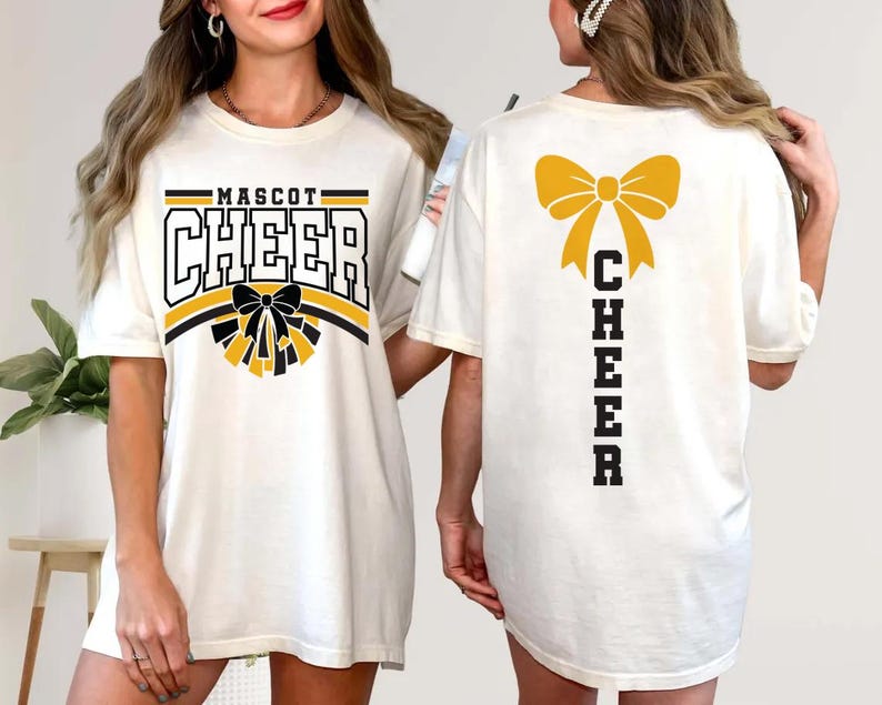 May include: Cream-colored t-shirt with the word "MASCOT CHEER" in black and gold lettering. The front features a bow and pom-pom design. The back of the shirt displays a gold bow and the word "CHEER" vertically.