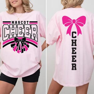 May include: Light pink t-shirt with "MASCOT CHEER" in black and white block letters, a black and pink bow, and pom-poms. The back of the shirt features a large pink bow and the word "CHEER" in black block letters.