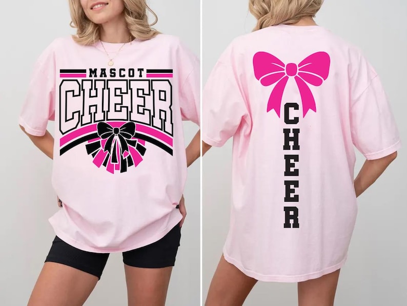 May include: Light pink t-shirt with "MASCOT CHEER" in black and white block letters, a black and pink bow, and pom-poms. The back of the shirt features a large pink bow and the word "CHEER" in black block letters.