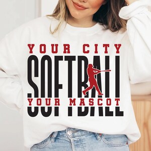 May include: White sweatshirt with the words "YOUR CITY SOFTBALL YOUR MASCOT" in black and red lettering. A red softball player graphic is in the center. The sweatshirt is a casual sports-themed apparel.