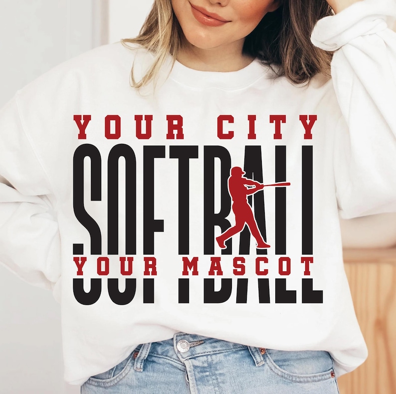 May include: White sweatshirt with the words "YOUR CITY SOFTBALL YOUR MASCOT" in black and red lettering. A red softball player graphic is in the center. The sweatshirt is a casual sports-themed apparel.