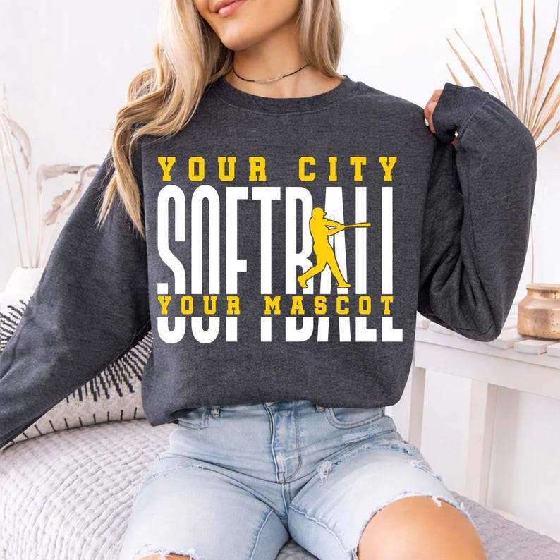 May include: Dark gray sweatshirt with the text "YOUR CITY SOFTBALL YOUR MASCOT" in white and yellow. A yellow softball player graphic is in the center. The sweatshirt is a casual style.