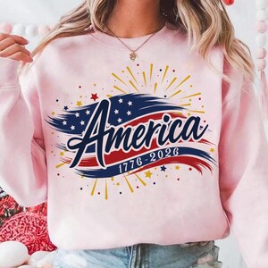 May include: A pale pink sweatshirt featuring the word "America" in a decorative font, with a design incorporating the colours of the American flag and stars. The text "1776-2026" is below. The design includes yellow and red bursts.