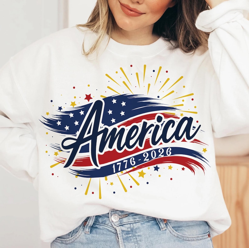 May include: White sweatshirt with a patriotic design. The design features the word "America" in a script font, with a red, white, and blue colour scheme. The design also includes the years "1776-2026" and star and firework accents.