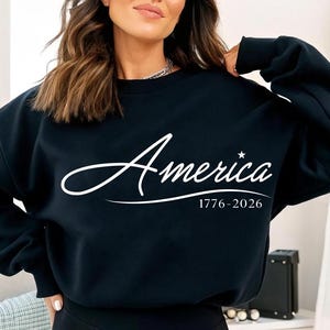 May include: A black sweatshirt with the word "America" in white script, a star, and the dates 1776-2026. The sweatshirt is a crew neck style.