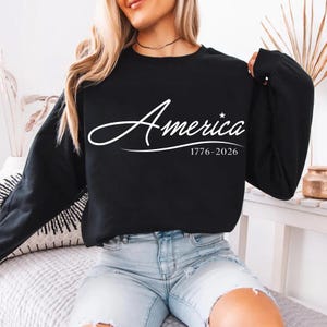 May include: Black crew neck sweatshirt with the word "America" in white script, a star, and the dates "1776-2026". The sweatshirt is a long-sleeved top.