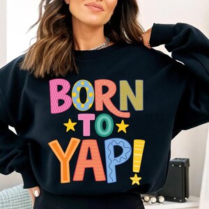 May include: Black sweatshirt with the text "BORN TO YAP!" in colorful, playful lettering. The design includes pink, orange, green, blue, and yellow, with star accents. A casual, fun graphic tee.
