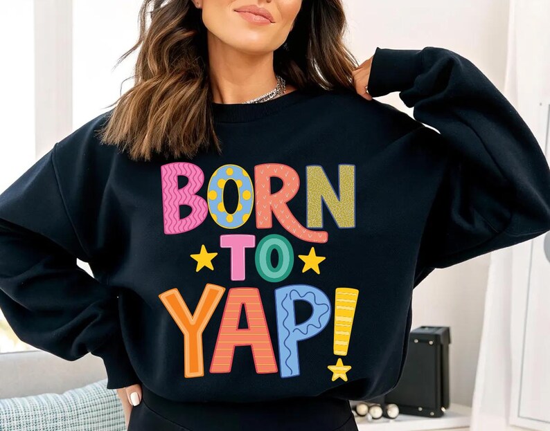 May include: Black sweatshirt with the text "BORN TO YAP!" in colorful, playful lettering. The design includes pink, orange, green, blue, and yellow, with star accents. A casual, fun graphic tee.