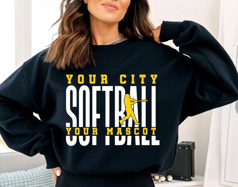 May include: Black sweatshirt with the text "YOUR CITY SOFTBALL YOUR MASCOT SOFTBALL" in white and yellow. A yellow softball player graphic is in the center. The sweatshirt is a casual style.