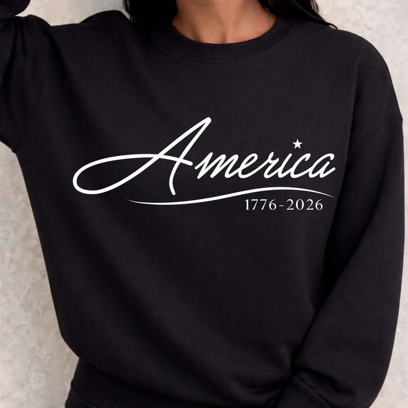 May include: Black sweatshirt with the word "America" in a white script font. Below the word is the text "1776-2026". A small white star is above the letter "a" in "America". The sweatshirt is a crew neck style.