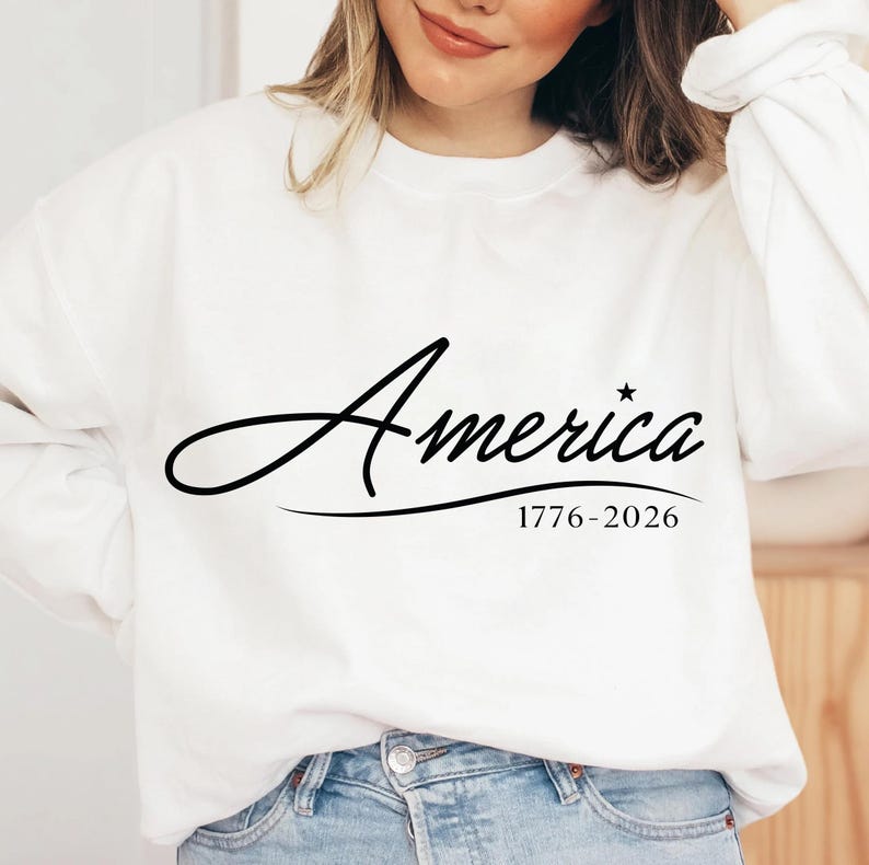 May include: A white sweatshirt with the word "America" in a cursive font, accompanied by a star and the dates "1776-2026" in black. The sweatshirt is a casual style.