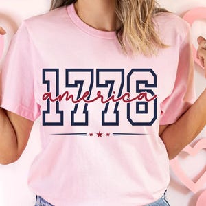 May include: A light pink short-sleeve t-shirt featuring the navy blue block-style text 1776 with the word america written in a red cursive script across the center. Below the text is a horizontal line with three small red stars in the middle.