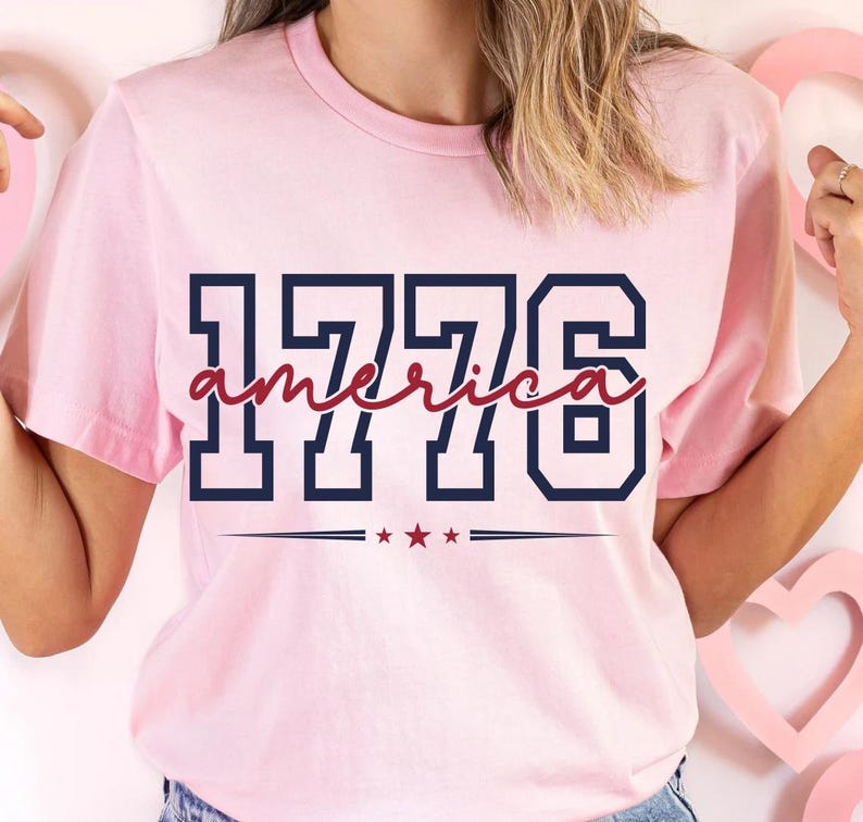 May include: A light pink short-sleeve t-shirt featuring the navy blue block-style text 1776 with the word america written in a red cursive script across the center. Below the text is a horizontal line with three small red stars in the middle.