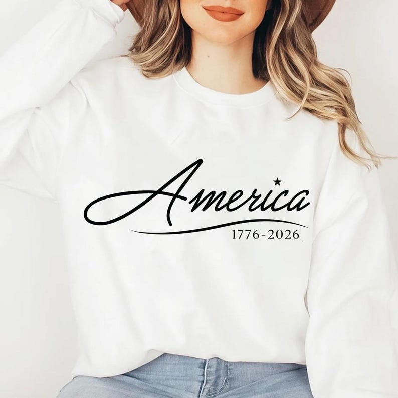 May include: White sweatshirt with the word "America" in a cursive font, accompanied by a star and the dates "1776-2026" in black. The sweatshirt is a crew neck style.