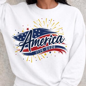 May include: White sweatshirt with a patriotic design. The graphic features the word "America" in a stylized font, with a blue and red colour scheme, and the dates "1776-2026". The design includes stars and fireworks.