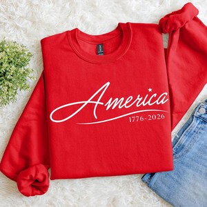 May include: A red crewneck sweatshirt with the word "America" in white script, along with the dates "1776-2026" and a small star. The sweatshirt is folded, with a portion of blue jeans visible.