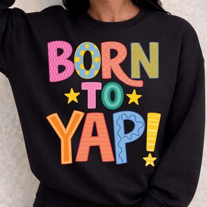 May include: Black sweatshirt with the words "BORN TO YAP" in colorful, block letters. The text includes pink, blue, yellow, orange, and green. Yellow stars are placed around the words. The sweatshirt is a crew neck.