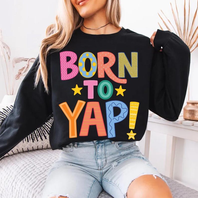 May include: Black sweatshirt with the text "BORN TO YAP!" in colorful, playful lettering. The design includes pink, blue, orange, and yellow text with star accents. A casual, comfortable top.