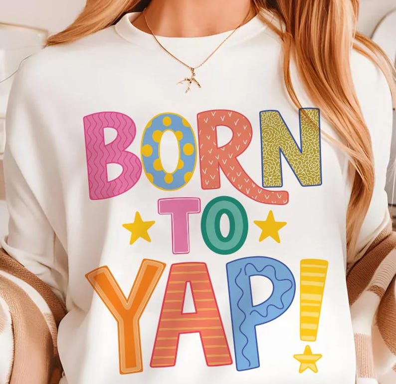 May include: White sweatshirt with the phrase "BORN TO YAP!" in colorful, playful lettering. The design features pink, blue, orange, and yellow text with star accents, creating a fun and vibrant look. A casual and comfortable top.