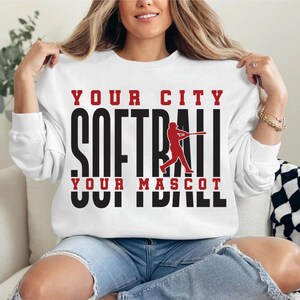 May include: White sweatshirt with the text "YOUR CITY SOFTBALL YOUR MASCOT" in black and red. A red silhouette of a softball player swinging a bat is in the center. The sweatshirt is long-sleeved.