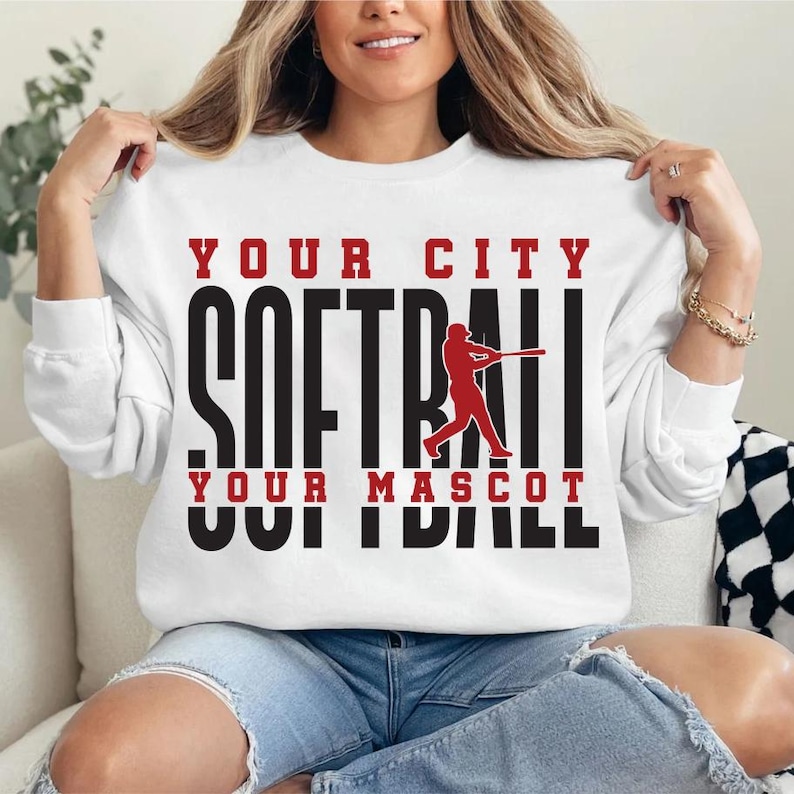 May include: White sweatshirt with the text "YOUR CITY SOFTBALL YOUR MASCOT" in black and red. A red silhouette of a softball player swinging a bat is in the center. The sweatshirt is long-sleeved.