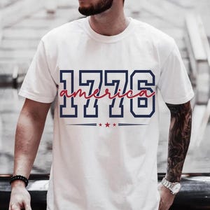 May include: A white short-sleeved t-shirt featuring the navy blue block-style text 1776 with the word america written in a red cursive script across the center. Below the text are three small red stars centered between two horizontal navy lines.