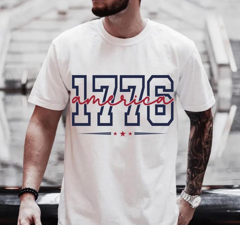 May include: A white short-sleeved t-shirt featuring the navy blue block-style text 1776 with the word america written in a red cursive script across the center. Below the text are three small red stars centered between two horizontal navy lines.