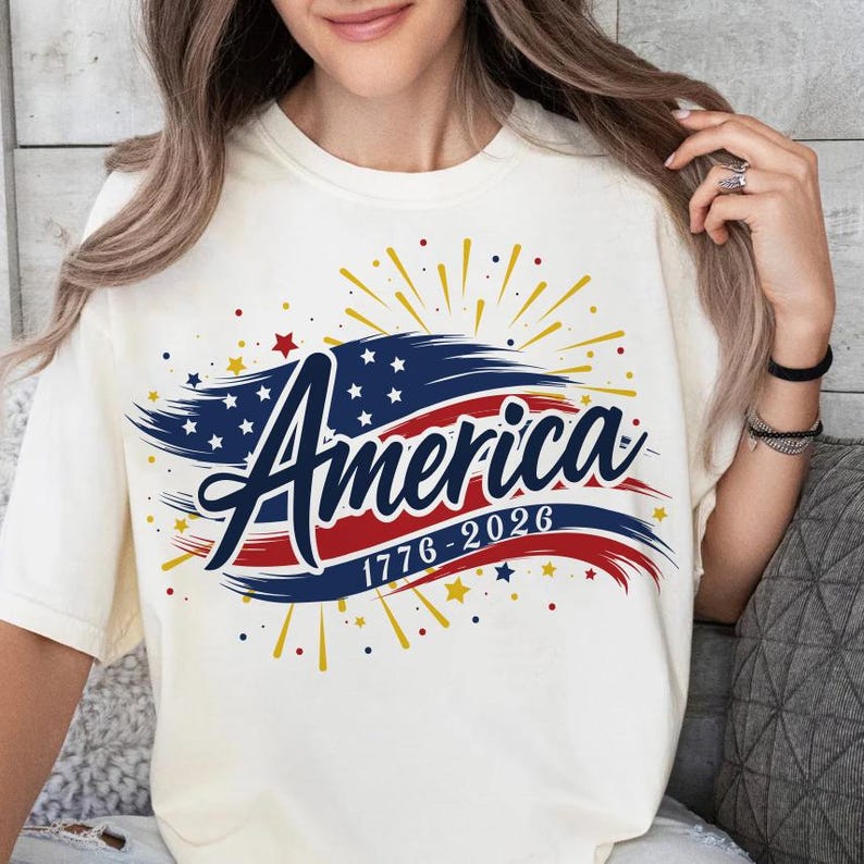 May include: Off-white t-shirt with the word "America" in a script font, set against a blue and red banner design. The dates "1776-2026" are below. The design includes stars and yellow and gold bursts.