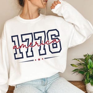 May include: A white long-sleeve sweatshirt featuring the navy blue block-style numbers 1776. The word america is written in red cursive script across the numbers, with three small red stars and horizontal lines positioned below the text.