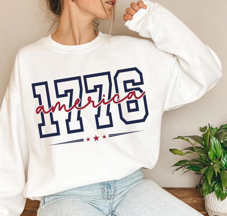 May include: A white long-sleeve sweatshirt featuring the navy blue block-style numbers 1776. The word america is written in red cursive script across the numbers, with three small red stars and horizontal lines positioned below the text.