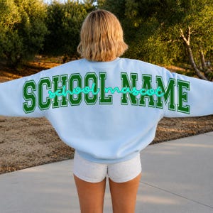 May include: A light blue crewneck sweatshirt featuring the text SCHOOL NAME in large green block letters across the back, with the word school mascot written in a smaller, cursive, bright turquoise script font layered over the green text.