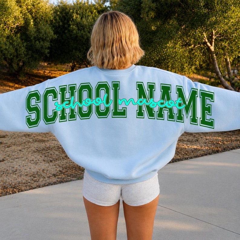 May include: A light blue crewneck sweatshirt featuring the text SCHOOL NAME in large green block letters across the back, with the word school mascot written in a smaller, cursive, bright turquoise script font layered over the green text.