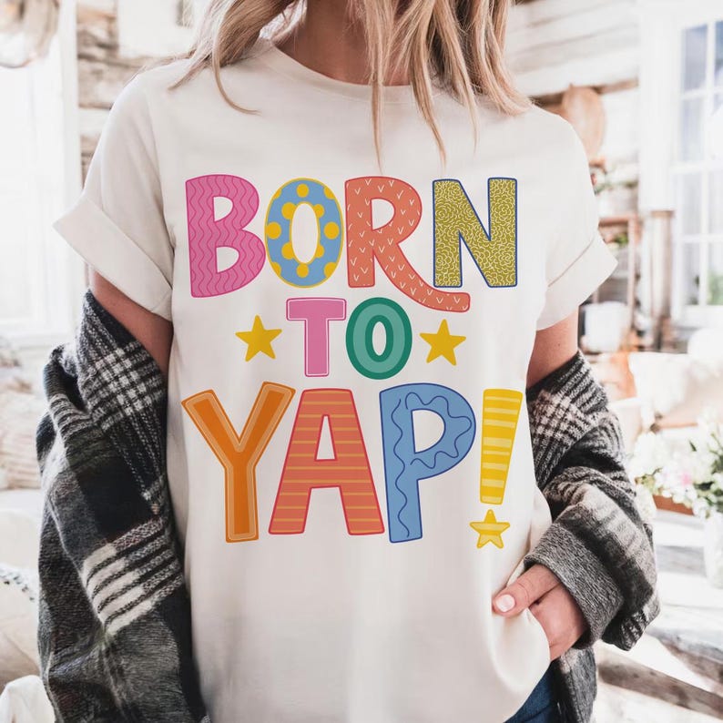 May include: Cream-colored t-shirt with the phrase "BORN TO YAP!" in colorful, playful lettering. The text features various patterns and colors, including pink, yellow, orange, and blue. Two yellow stars flank the word "TO".