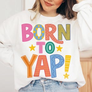 May include: White sweatshirt with the phrase "BORN TO YAP!" in colorful, patterned letters. The text includes pink, blue, orange, yellow, and green. The design features stars and exclamation points.