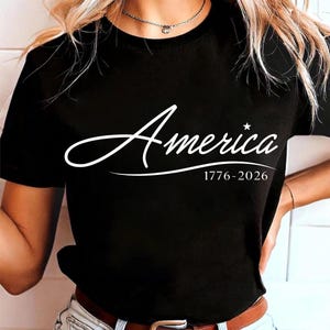 May include: Black t-shirt with the word "America" in a white script font, accompanied by the dates "1776-2026" below. The design features a star above the "a" in "America".