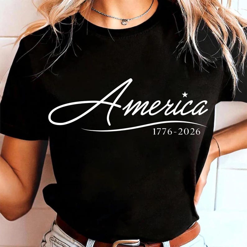 May include: Black t-shirt with the word "America" in a white script font, accompanied by the dates "1776-2026" below. The design features a star above the "a" in "America".