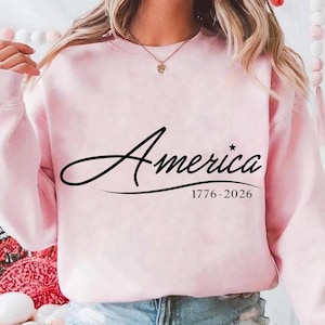May include: A light pink sweatshirt with the word "America" in a black script font. Below the word is the text "1776-2026". The sweatshirt is a solid color and has long sleeves.