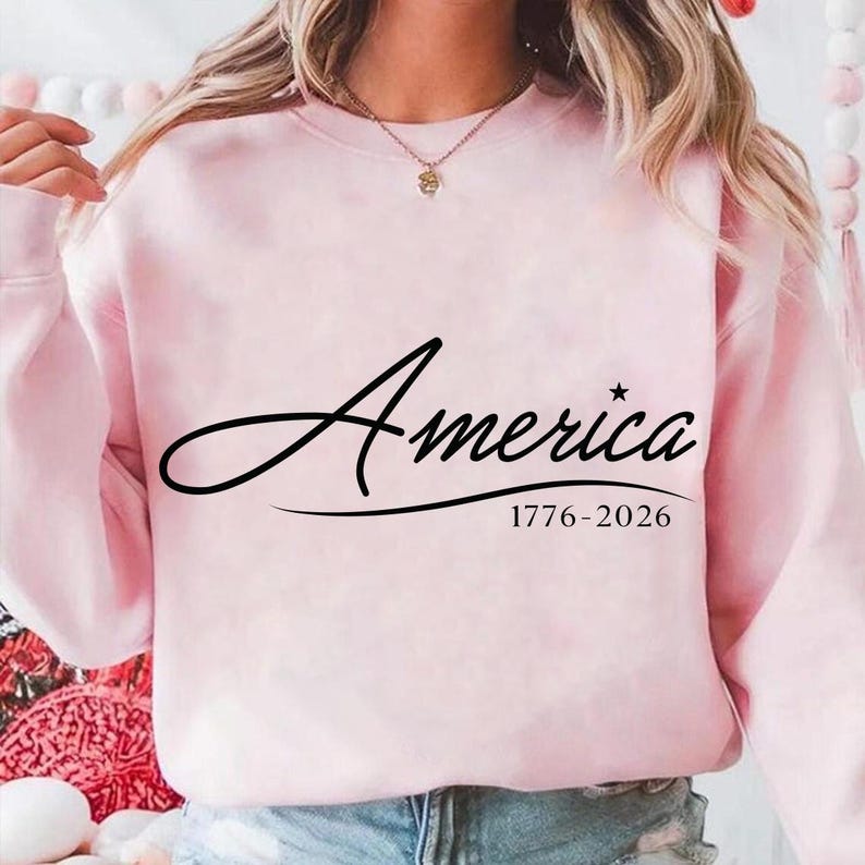 May include: A light pink sweatshirt with the word "America" in a black script font. Below the word is the text "1776-2026". The sweatshirt is a solid color and has long sleeves.