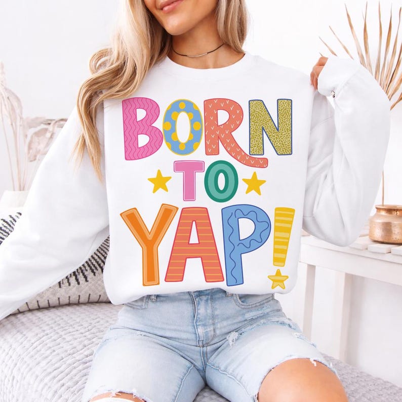 May include: White sweatshirt with the text "BORN TO YAP!" in colorful, playful lettering. The design features pink, orange, yellow, green, and blue letters with star accents. A casual and fun graphic top.
