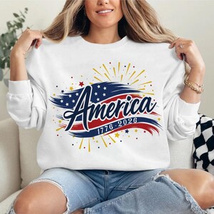 May include: White sweatshirt featuring the word "America" in a patriotic design. The design incorporates a blue and red flag motif, stars, and the years "1776-2026". The sweatshirt has long sleeves.