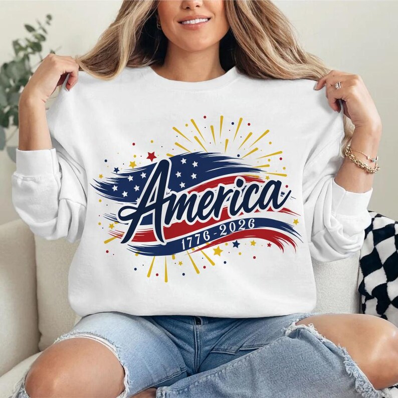 May include: White sweatshirt featuring the word "America" in a patriotic design. The design incorporates a blue and red flag motif, stars, and the years "1776-2026". The sweatshirt has long sleeves.